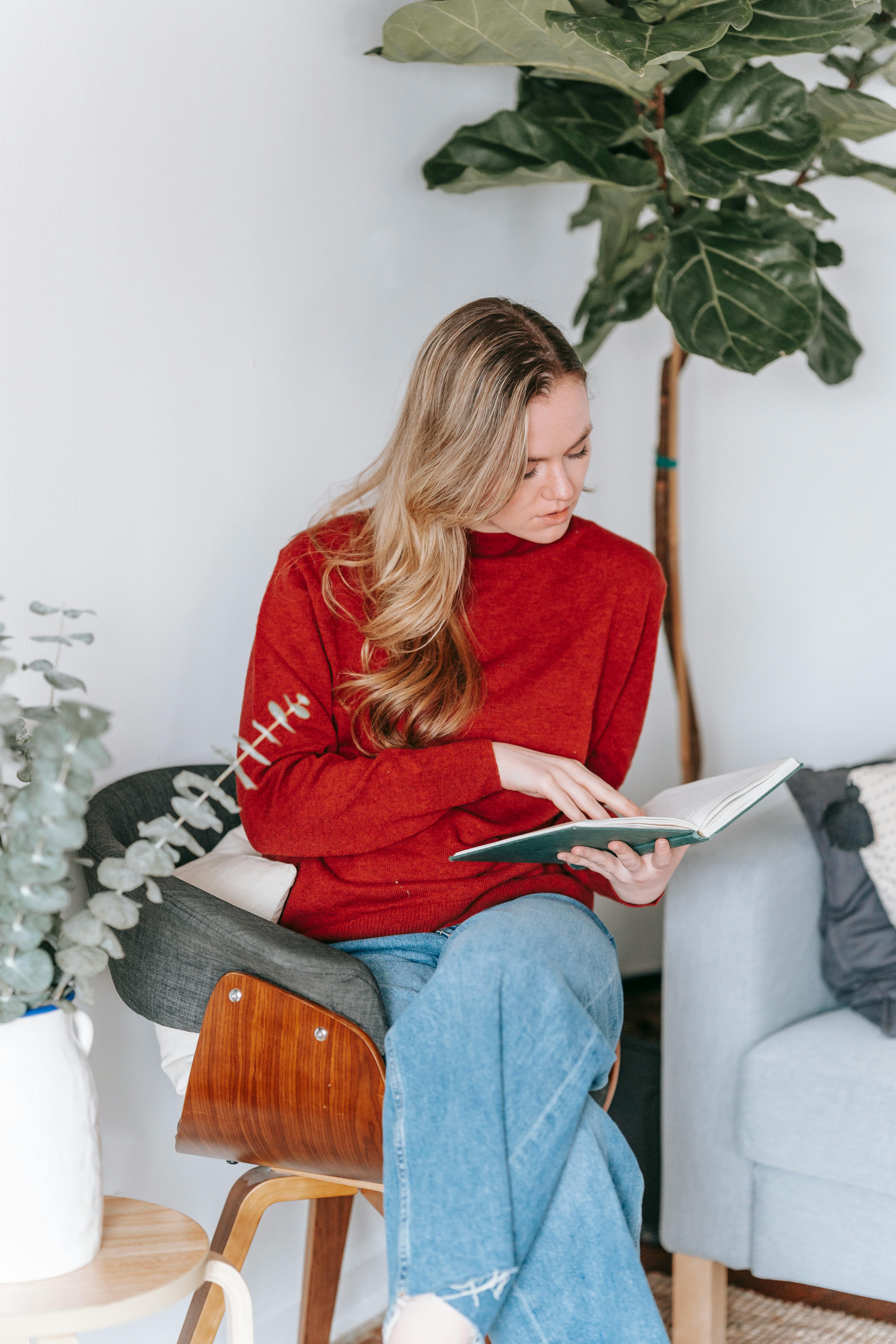 thoughtful young woman reading textbook on chair at home