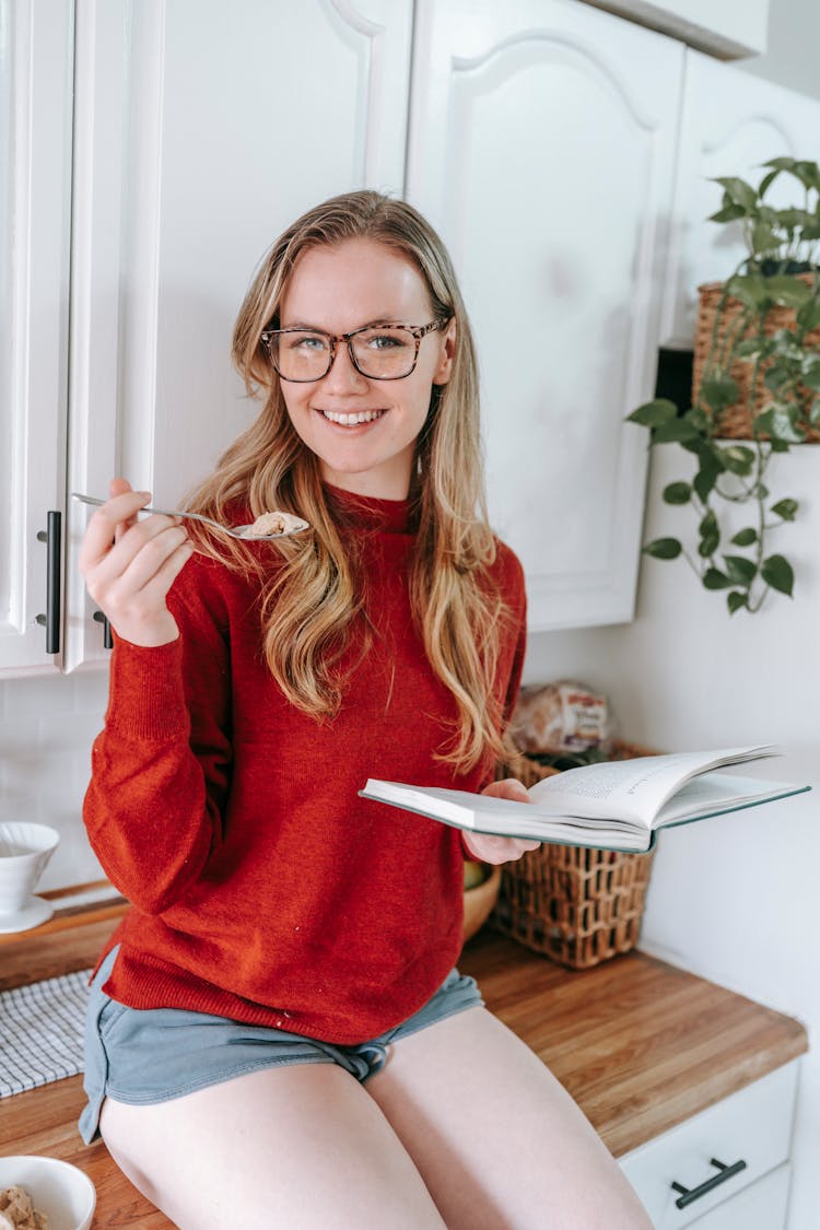 Smiling Lady Eating And Reading Book Sitting On Cabinet In Kitchen