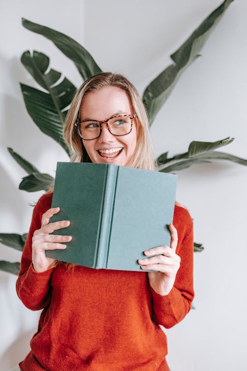 Joyful young female student with blond hair in sweater and eyeglasses smiling and looking away while standing near white wall with book in hands after success in exam
