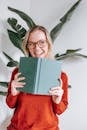 Joyful young female student with blond hair in sweater and eyeglasses smiling and looking away while standing near white wall with book in hands after success in exam