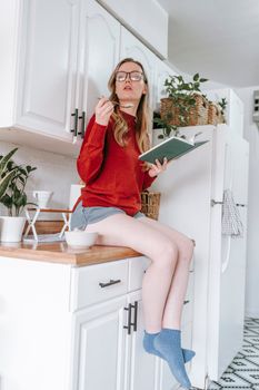 Full body of young smart lady in casual clothes and eyeglasses sitting on kitchen cabinet and eating yogurt while reading book in light apartment