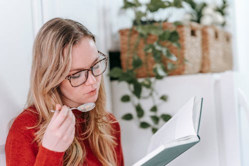 Free Thoughtful young female millennial with long blond hair in casual clothes and eyeglasses eating yummy food and reading interesting book during breakfast in light kitchen Stock Photo