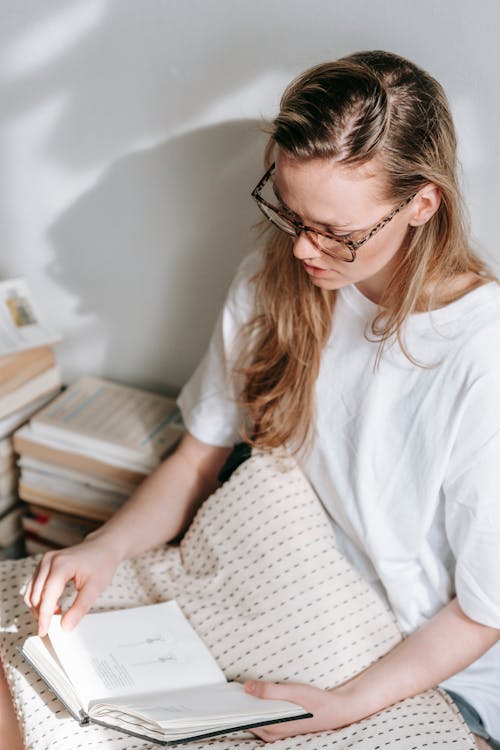 From above of serious young lady with long hair in white t shirt and eyeglasses sitting on floor with pillow near stack of books and reading interesting novel in daylight