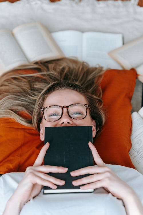 Free From above of positive young female in eyeglasses covering face with book while lying on cushion and looking at camera Stock Photo