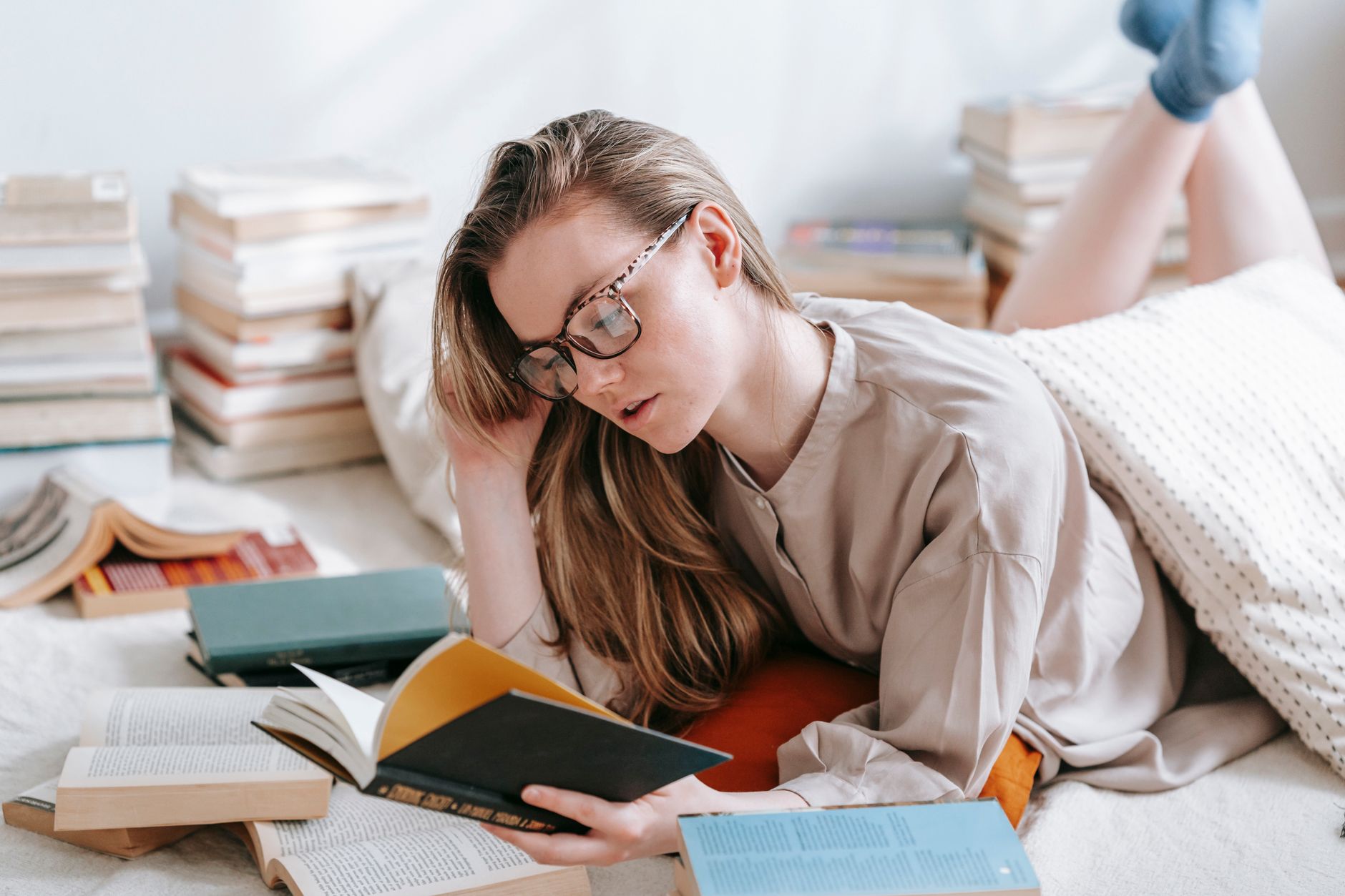 Concentrated female in eyeglasses reading book while lying on floor with plaid and collection of literature in light room at home