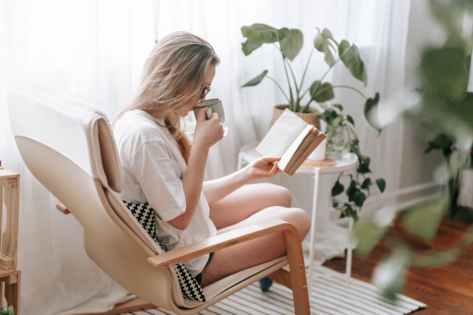 Side view of focused female drinking hot beverage while reading book in armchair in light room with potted green plants