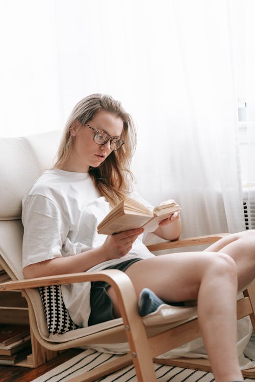 Free Serious female in casual clothes and eyeglasses reading book while sitting in armchair near window in light room at home Stock Photo