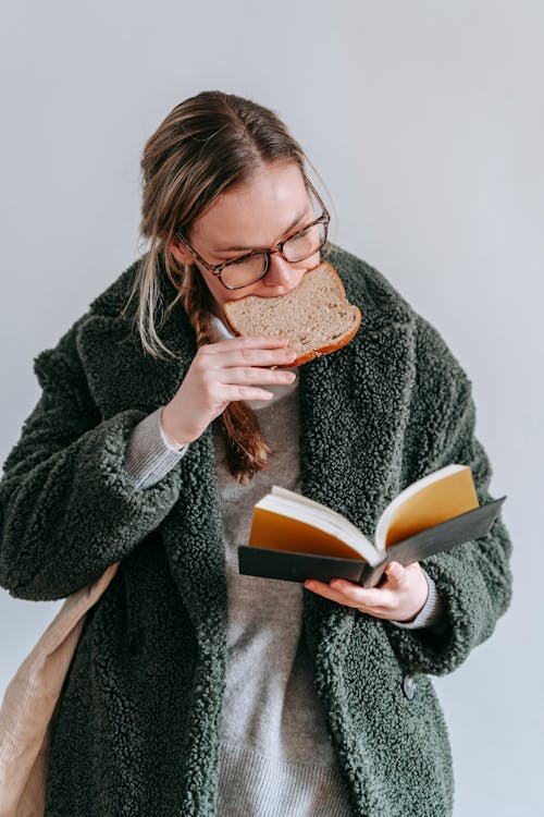 Concentrated female wearing warm coat and eyeglasses eating tasty bread while reading interesting book on white background in light room