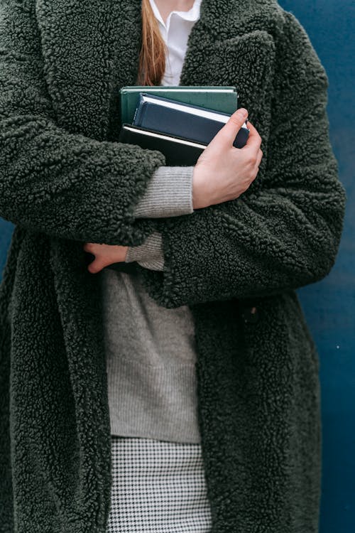Crop unrecognizable female in stylish warm outerwear with collection of books in hands standing near wall on street in city