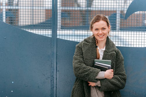 Cheerful female in warm outerwear with books in hands smiling and looking at camera while standing near metal fence on street