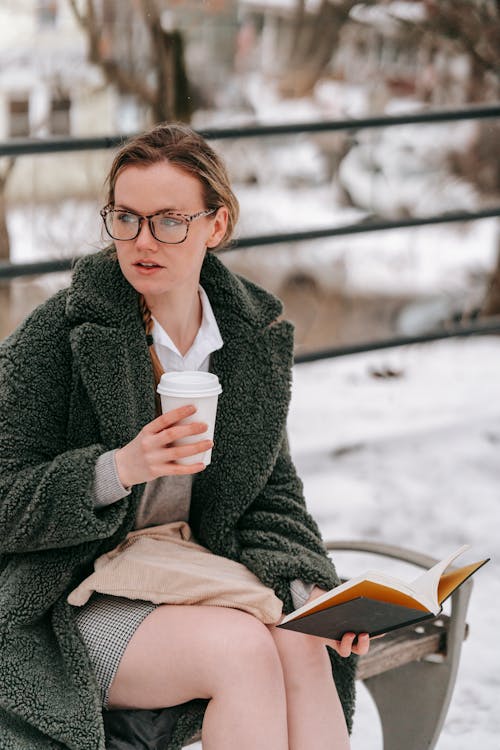 Serious female with paper cup of hot drink looking away and reading book while sitting on bench in winter time