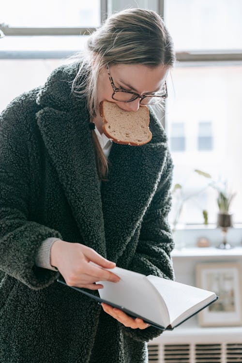 Concentrated young female in warm outerwear and eyeglasses flipping pages of book while eating bread in light room at home