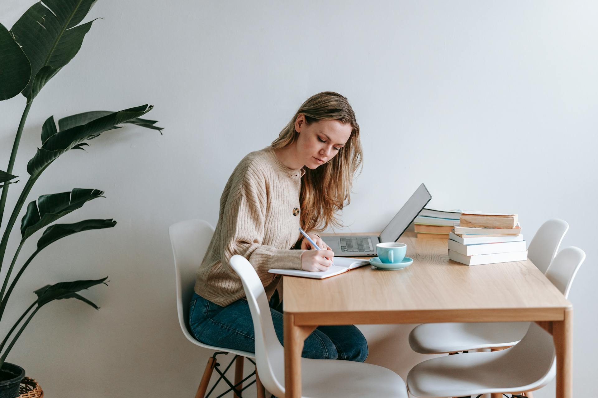 Side view of concentrated female freelancer taking notes in notepad while working at table with netbook in light room with green plant