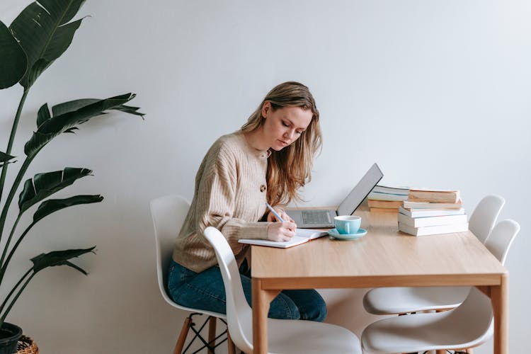 Focused Woman Writing In Notebook At Table With Laptop