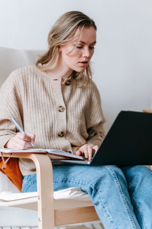 Free Serious female freelancer taking notes in notepad while looking at screen of netbook while sitting in armchair on white background Stock Photo