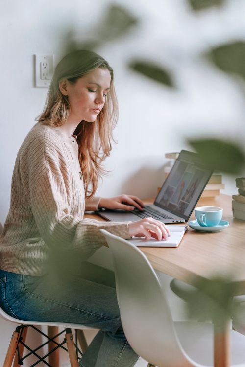 Free Side view of focused female freelancer reading notes in notepad while browsing netbook at table in room with blurred green leaves Stock Photo