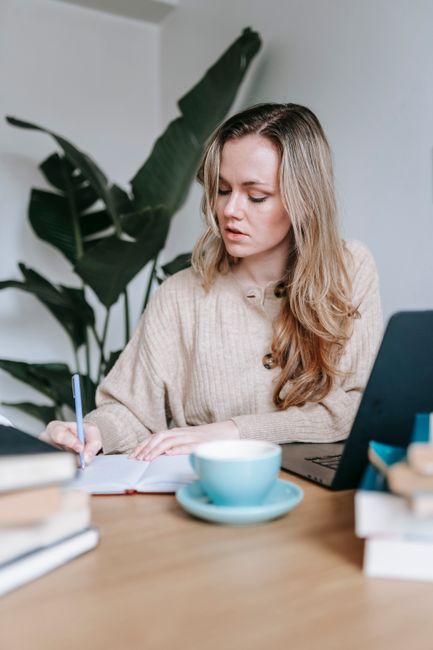 Thoughtful female freelancer taking notes in notepad while working at table with laptop at books near cup of hot drink