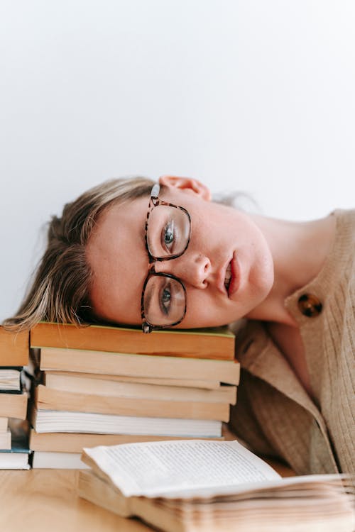 Calm young female in eyeglasses looking at camera while lying on stack of books on white background in light room