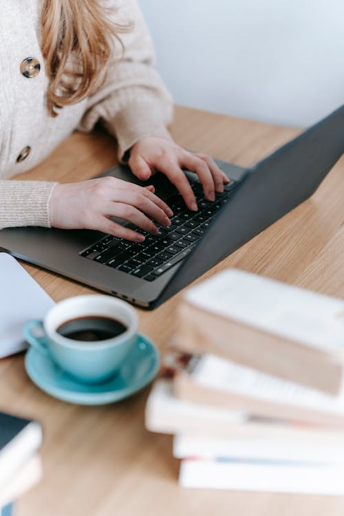 Free Crop anonymous female freelancer surfing netbook while sitting at wooden table with cup of hot coffee and books in room Stock Photo