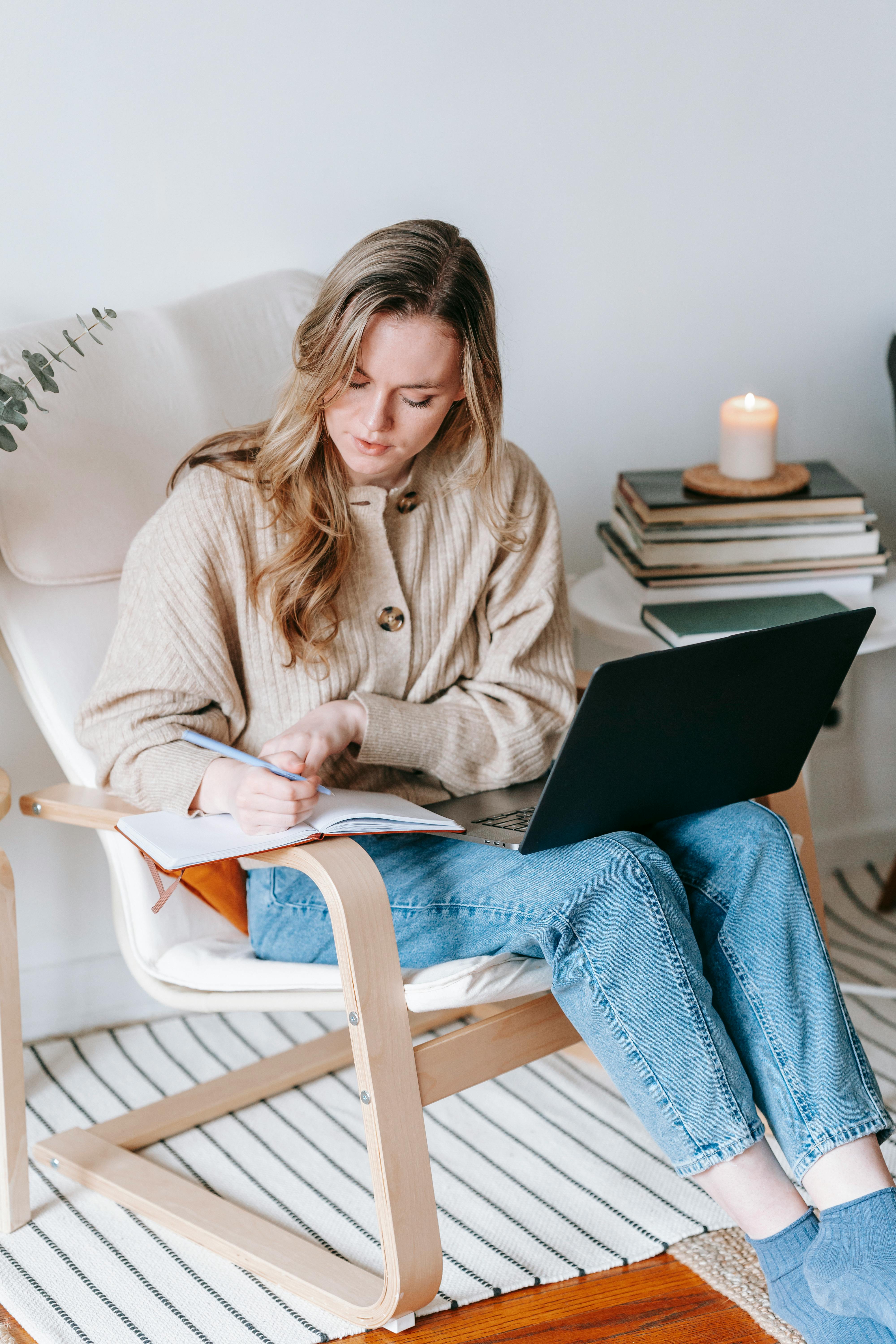 serious woman with laptop writing in notebook in armchair