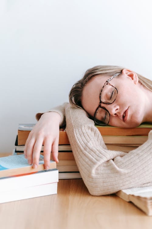 Tired female in eyeglasses sleeping at wooden table while lying on stack of books on white background in light room