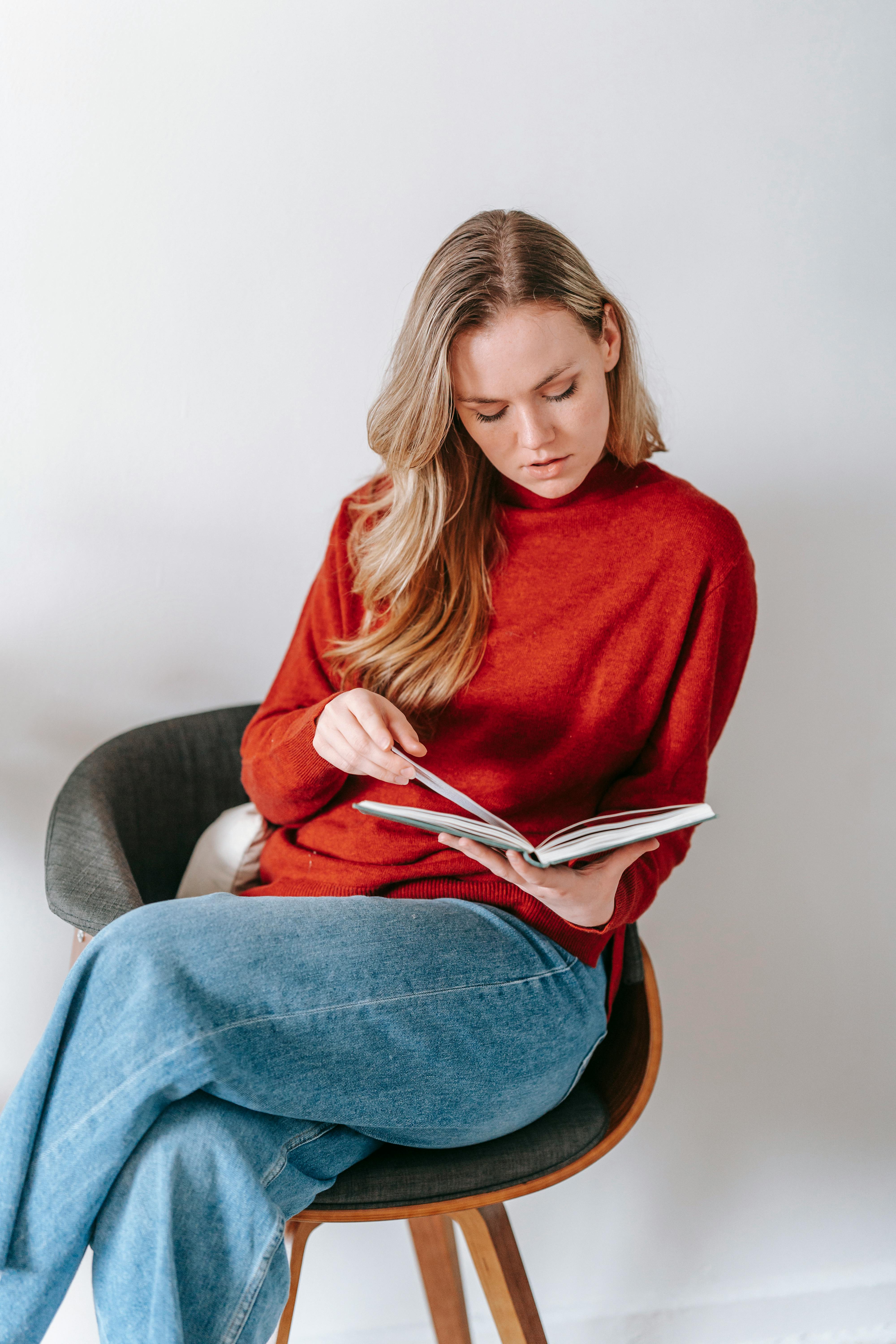 a woman in red sweater sitting on the chair while reading a book with her legs crossed