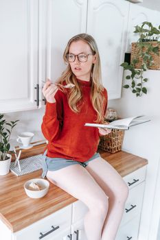 Blonde woman in casual wear sitting on kitchen counter, reading book and enjoying ice cream.