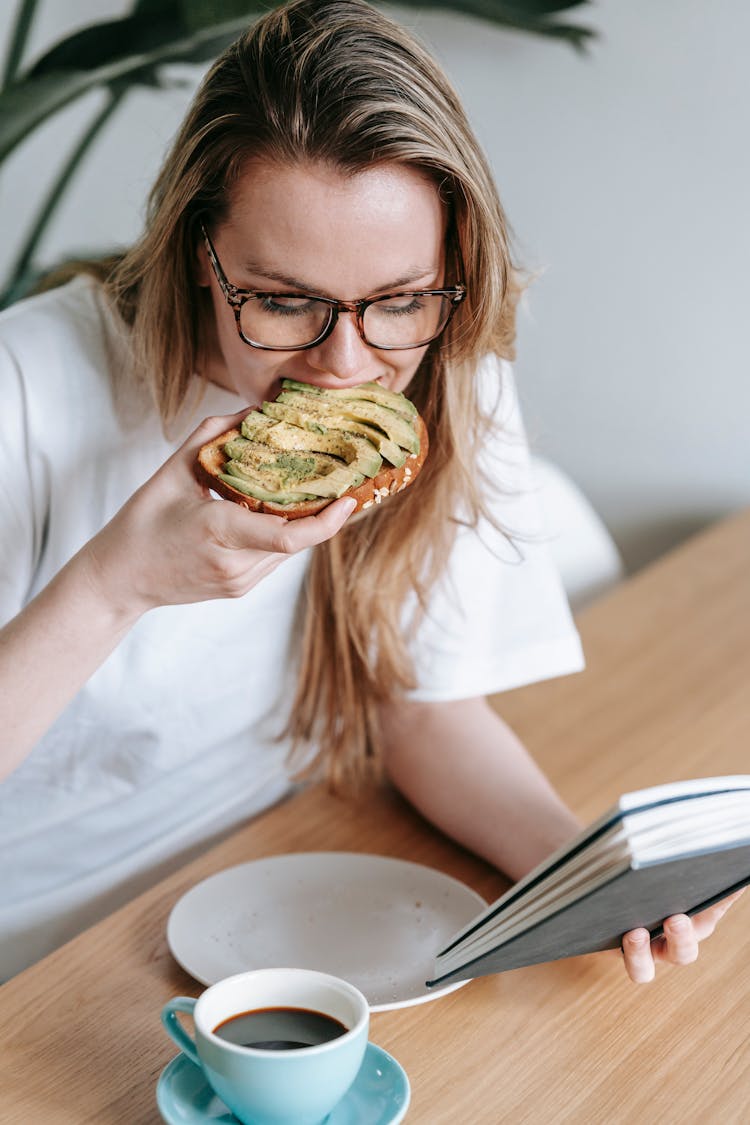 A Woman Eating Avocado Toast And Reading A Book