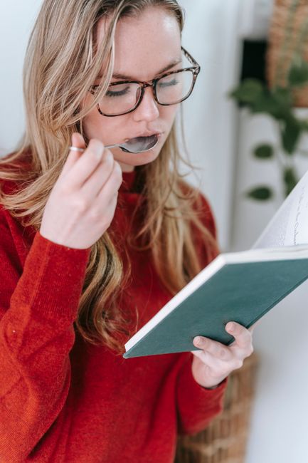 A woman enjoys reading a book and eating, dressed in a cozy red sweater, indoors.
