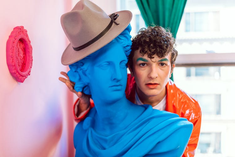 Man With Green Eyebrows In An Orange Jacket Near A Blue Statue