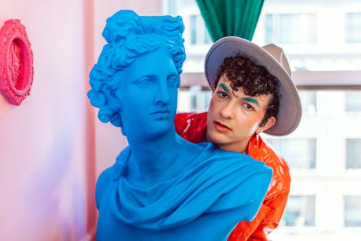 Vivid indoor portrait of a young man in a hat posing with a bright blue bust and playful makeup.