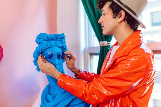 Stylish man in orange jacket and hat puts sunglasses on a vibrant blue bust indoors.