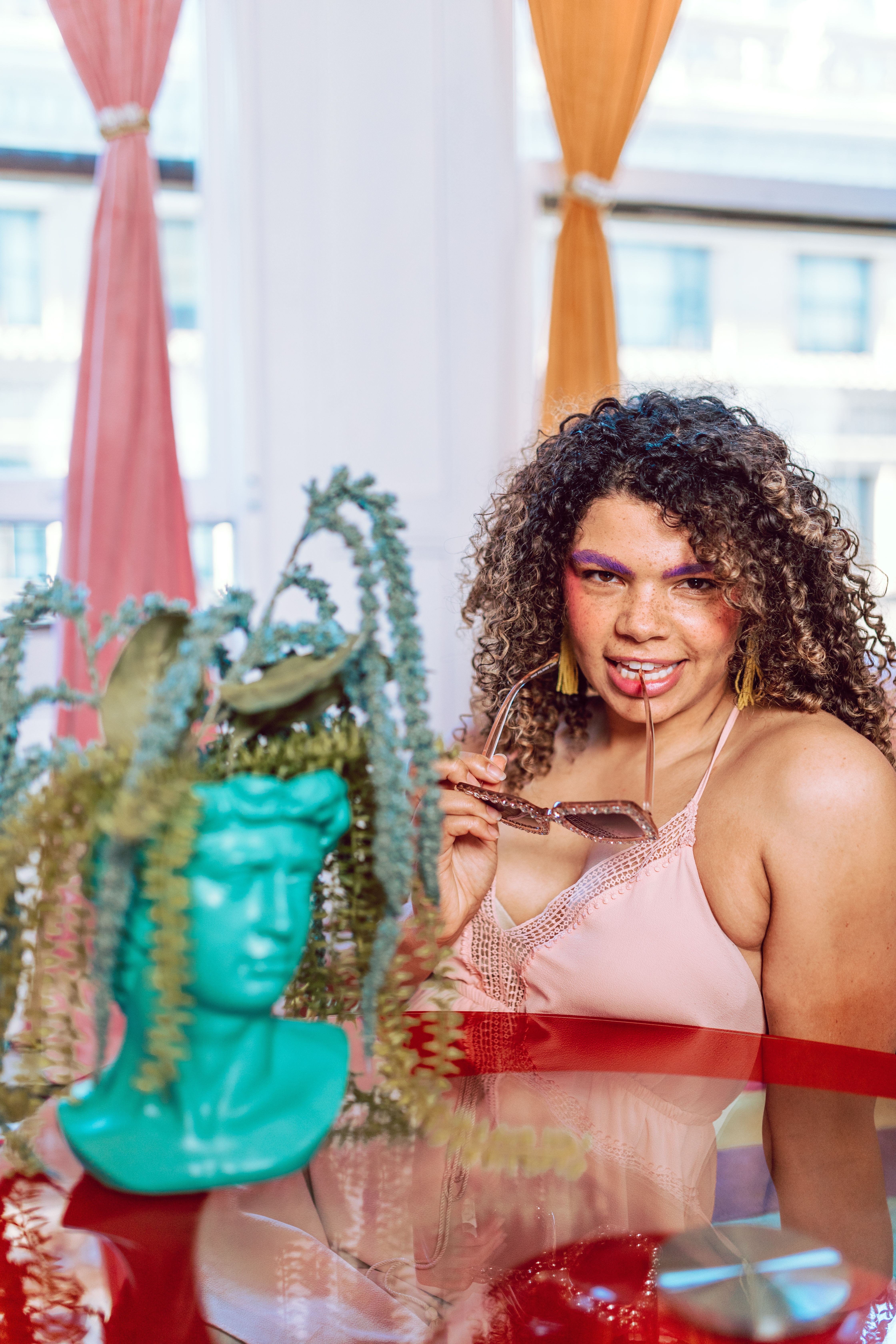 Fashionable woman with curly hair and sunglasses poses indoors near a decorative plant.