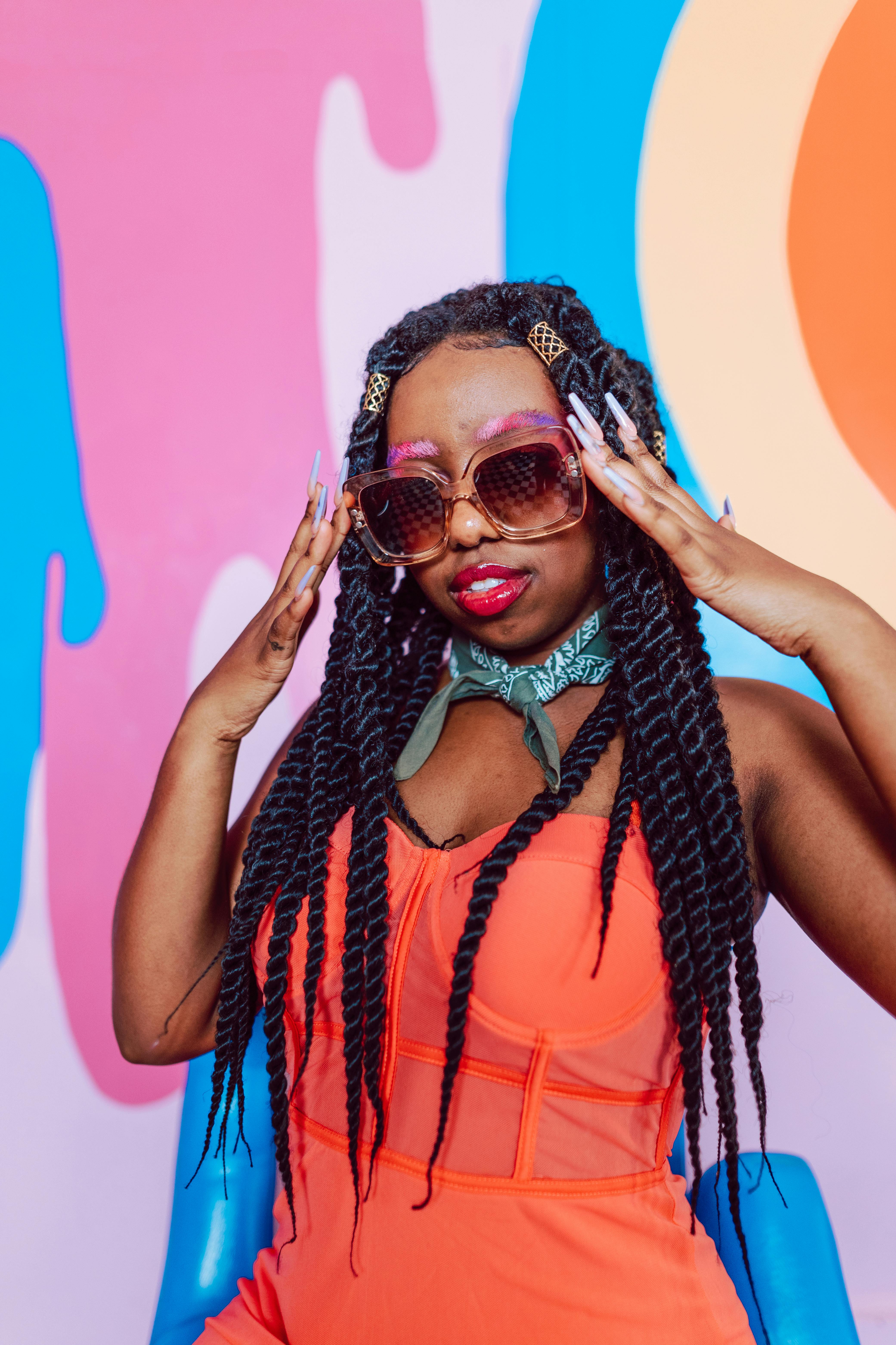 Stylish woman in an orange dress with sunglasses and braids, posing in front of a bright abstract mural.