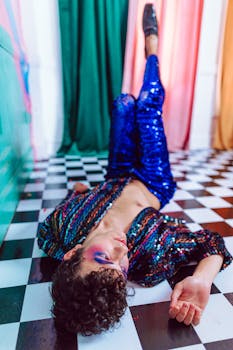 A vibrant person in sequined outfit lying on checkered floor with bold makeup.