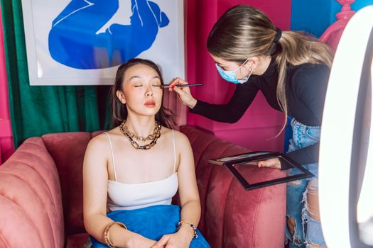 A makeup artist skillfully applies eyeshadow to a young woman sitting on a plush couch.