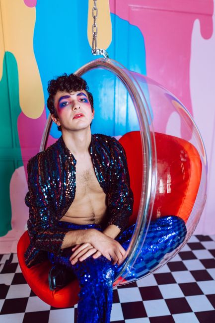 Colorful portrait of a young person in vibrant, contemporary fashion and makeup, sitting in a modern, artistic setting.