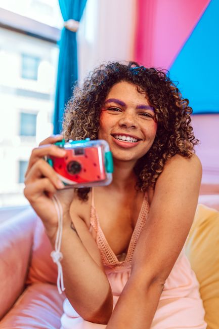 Cheerful woman with curly hair holds a camera, sitting in a colorful indoor setting.