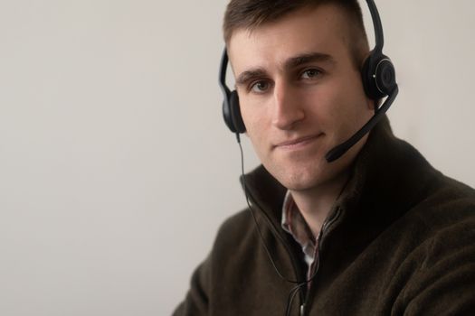 Positive young guy in casual outfit working on headphones with microphone in light workplace while looking at camera