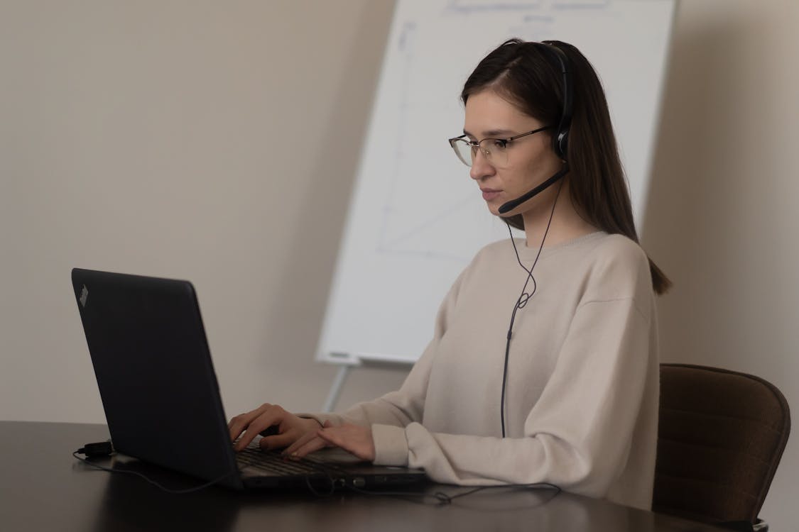 Free Serious young woman in casual outfit with eyeglasses sitting on chair at table and working on laptop with headphones with microphone near whiteboard in bright workplace Stock Photo