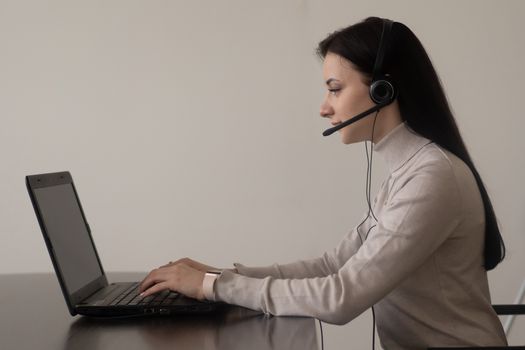 Side view of young female operator in casual clothes sitting on chair at table while working on laptop with headphones with microphone in light workplace