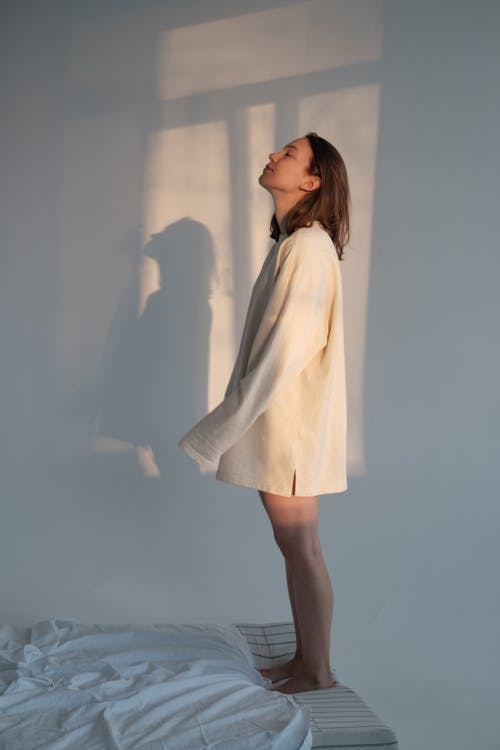 Free Pensive female in long sweater standing on bed in light room Stock Photo