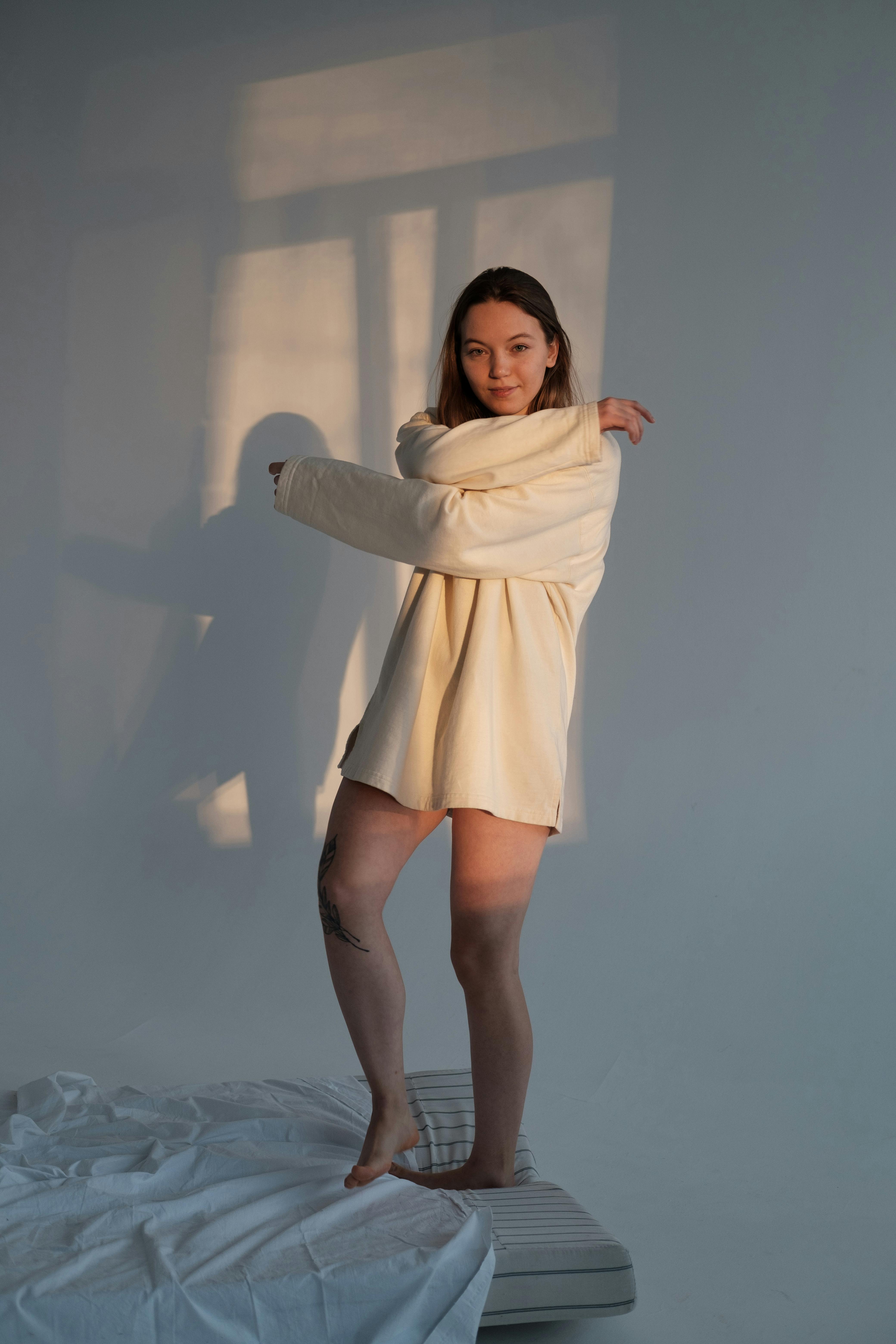 Can young female with brown hair in long sweater standing with crossed arms on beg against white wall in room in sunny day and looking at camera
