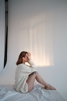 Side view of unrecognizable barefoot female touching face while sitting on soft mattress against white background