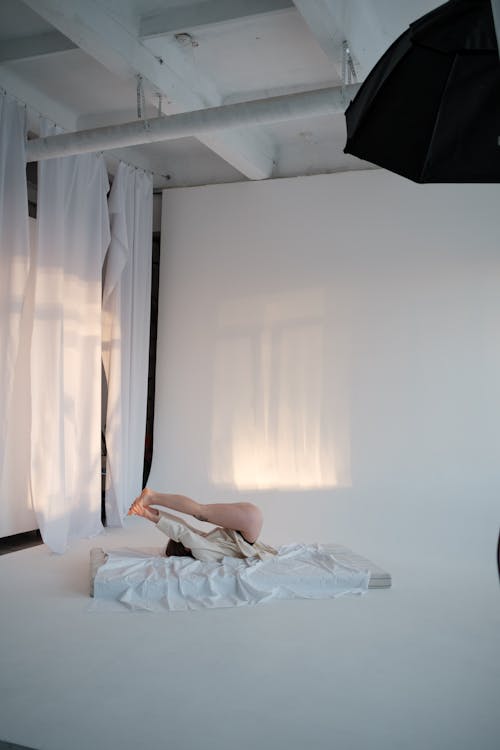 Side view of anonymous sensual female touching feet and rolling on back on soft mattress in light room