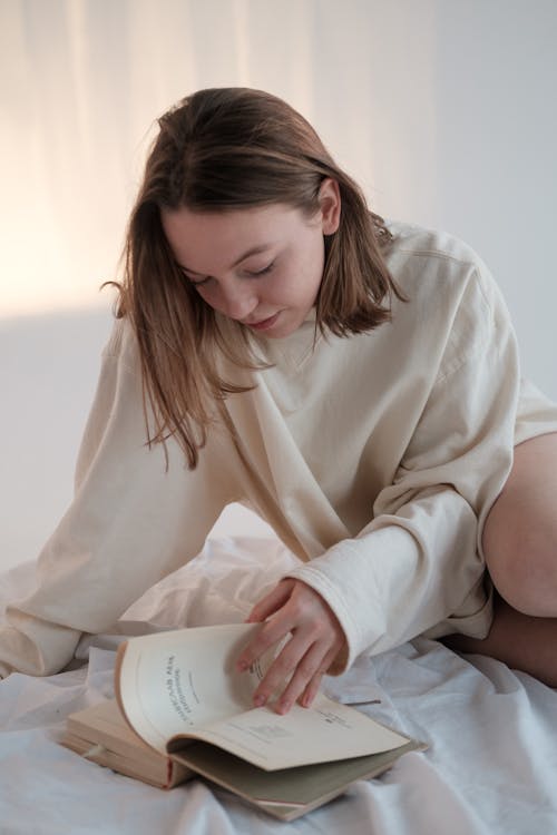 Free Young woman in sweatshirt flipping pages while sitting on bed and reading book in morning Stock Photo