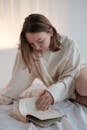 Young woman in sweatshirt flipping pages while sitting on bed and reading book in morning