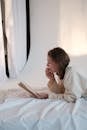 Side view of young woman touching face and reading interesting story while relaxing on mattress in morning
