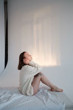 Side view of young barefoot woman in sweatshirt touching hair while sitting on mattress with closed eyes against white background