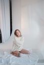 Young mad female in white straitjacket looking at camera while sitting still on mattress in light room